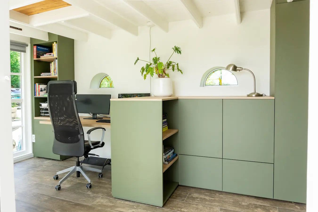 Thuiswerkplek met lichtgroene kasten, bureaustoel, computer en een plant bij kleine ramen onder een witte balkenplafond.