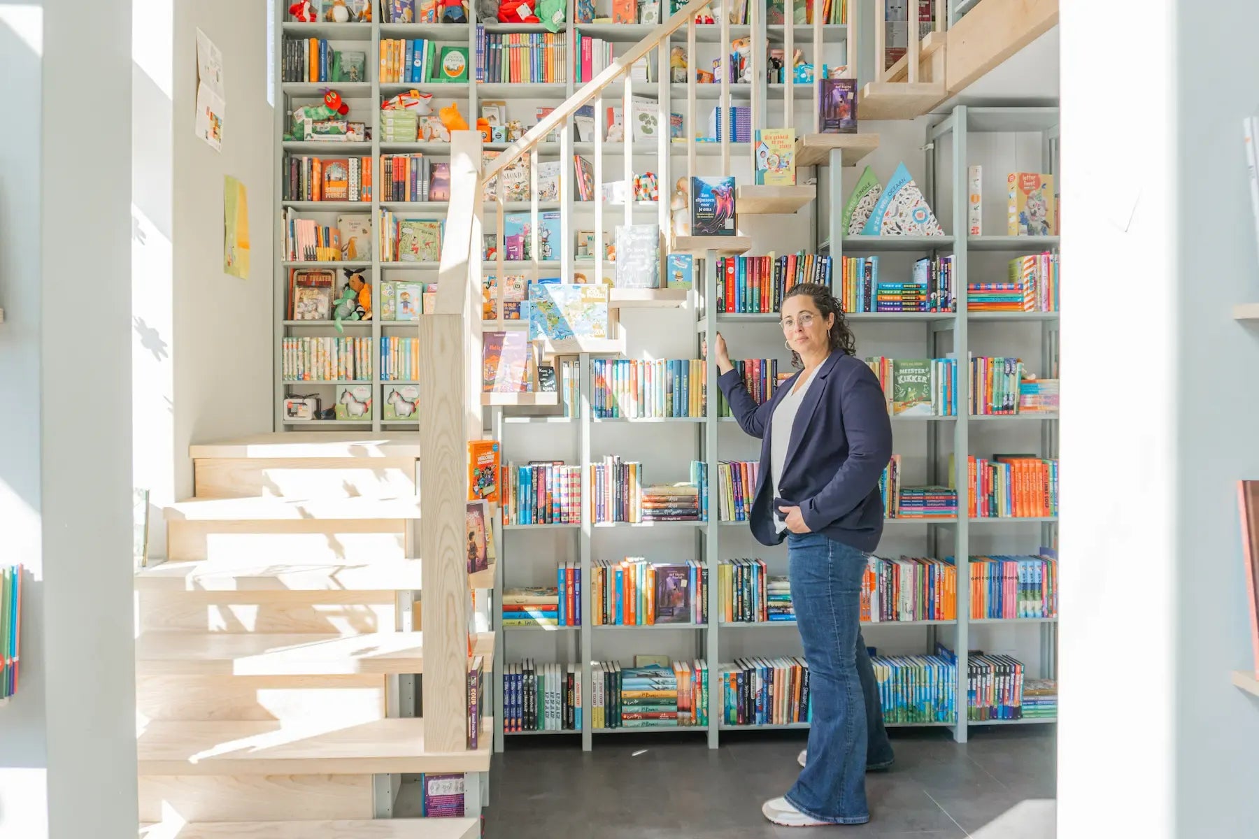 Vrouw staat bij hoge witte boekenkast vol kleurrijke boeken naast lichte houten trap in zonnige ruimte