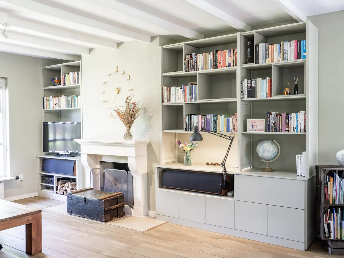 Lichtgrijze wandkast met open vakken gevuld met boeken, bureau met lamp en decoratie naast klassieke open haard in lichte ruimte