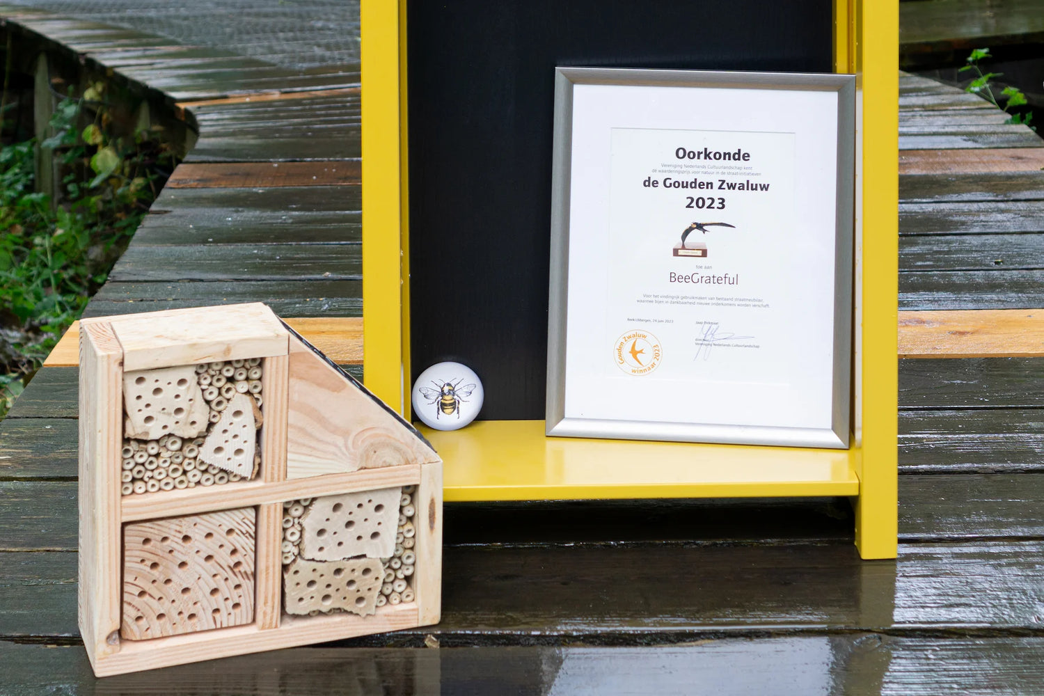 Houten bijenhotel naast bekroonde oorkonde met gele lijst op nat houten terraspad in bosrijke omgeving