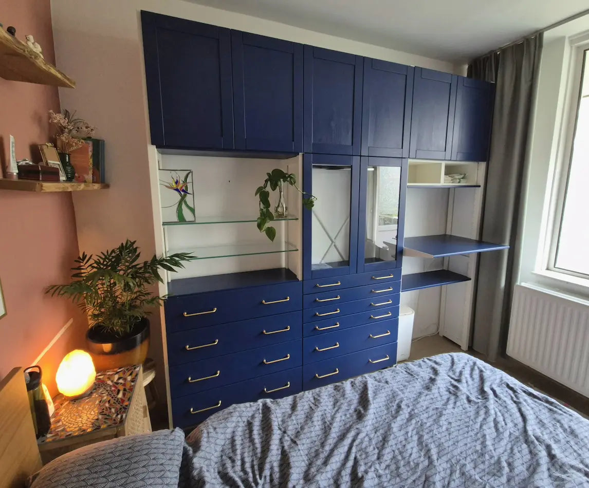 Matblauwe kastcombinatie met laden, vakken, glazen planken en open werkruimte naast een raam in slaapkamer.