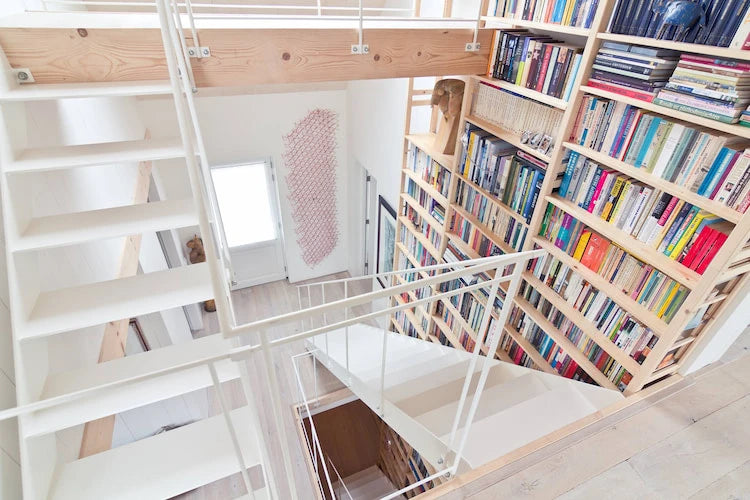 Moderne lichte trap met witte treden naast hoge houten boekenkast gevuld met kleurrijke boeken in open ruimte