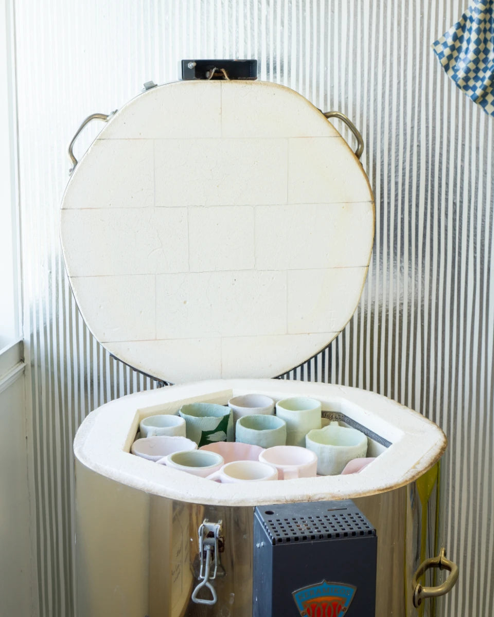 Open keramische oven met diverse pastelgekleurde keramiekpotten binnenin, naast raam en metalen wandpanelen