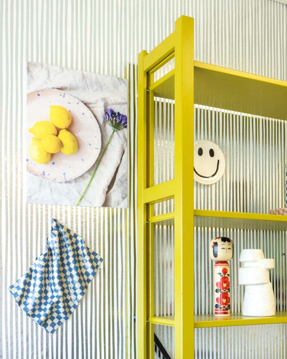 Felgeel open rek met keramische objecten en decoratief bord naast wand met foto van citroenen en blauwe bloemen