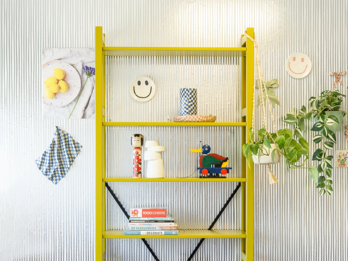 Geel open rek tegen geribbelde witte muur met keramische smileys, planten en kleurrijke decoratieve objecten