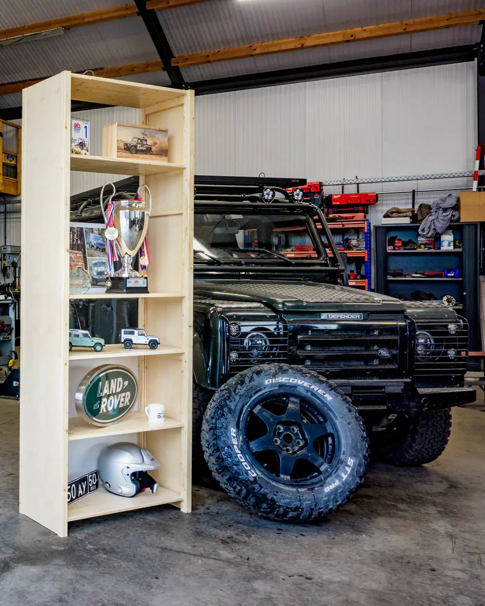 Houten open kast met schaalmodellen, trofeeën en helm naast zwarte Land Rover Defender en reserveband in garageomgeving