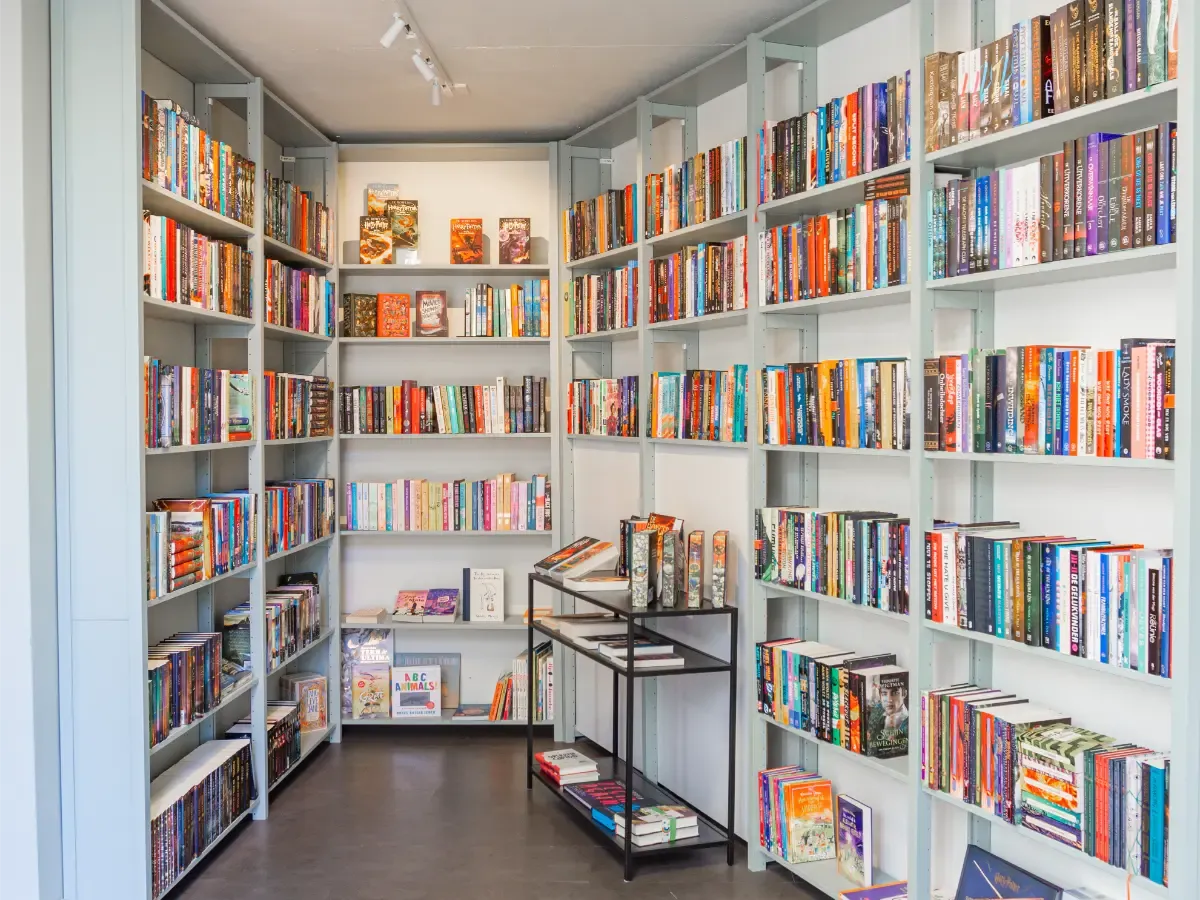 Witte boekenkasten vol diverse boeken in kleine ruimte met metalen rek en heldere verlichting vanaf boven