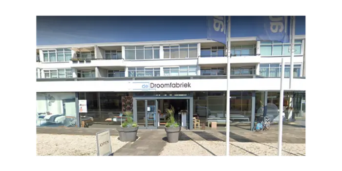 Moderne gevel van winkel De Droomfabriek met grote ramen, planten en drie vlaggenmasten ervoor bij zonnig weer.