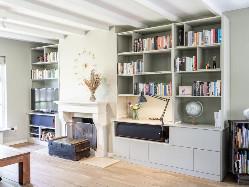 Lichte woonkamer met grijze wandkasten gevuld met boeken, open haard, houten vloer en sfeervolle werkplek met bureaulamp