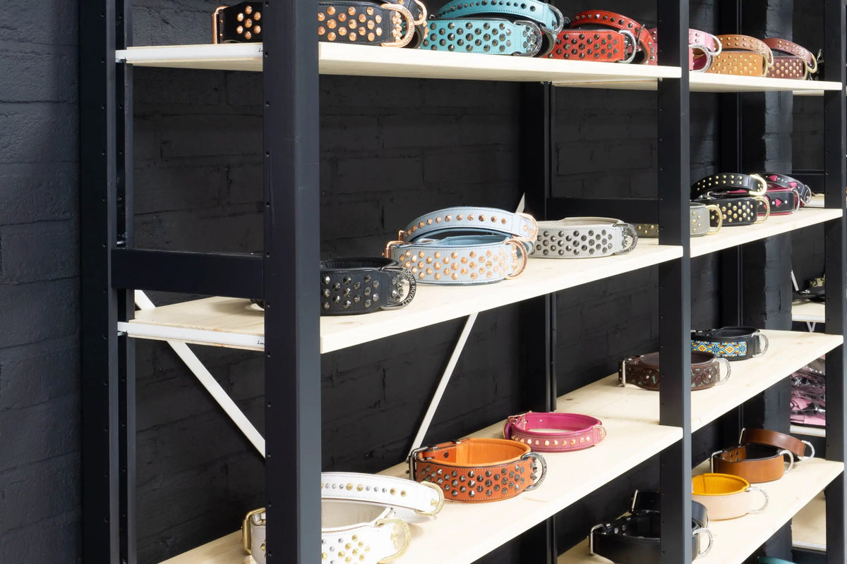 Zwart metalen rek met verschillende leren halsbanden in diverse kleuren en studdetails tegen zwarte bakstenen muur.